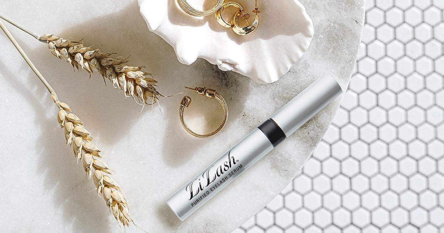LiLash Purified Eyelash Serum tube on marble with gold jewelry and wheat, beauty product flat lay