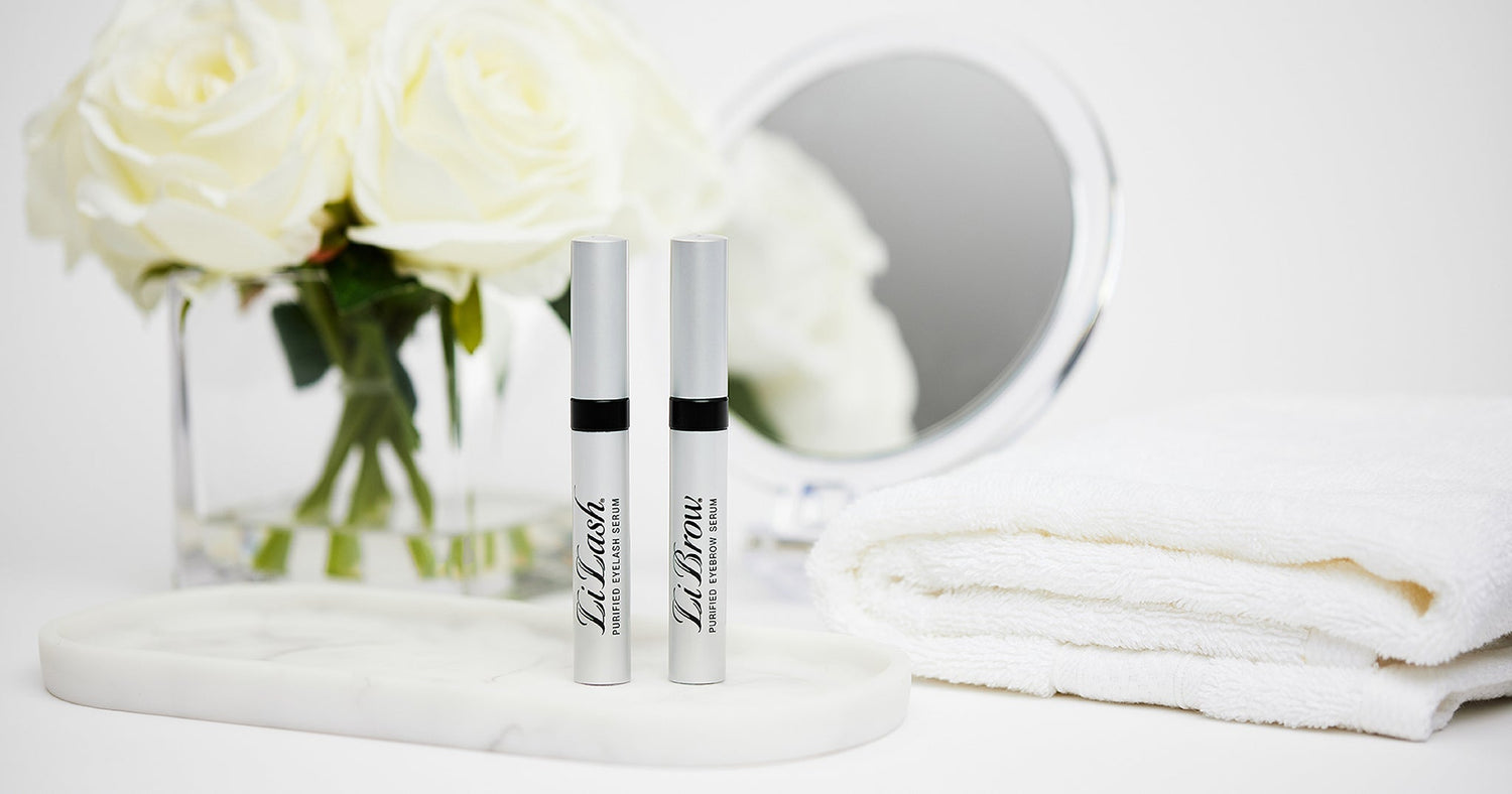 LiLash eyelash serum and LiBrow eyebrow serum on marble tray with white roses, mirror, and towel