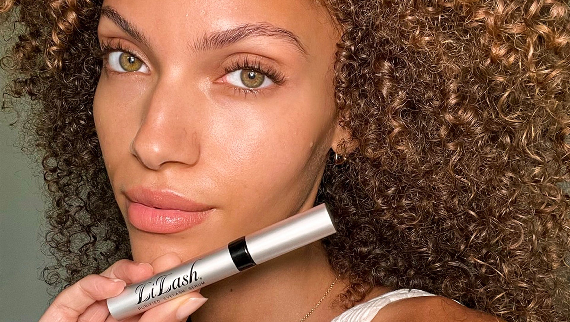 Woman with curly hair holding LiLash eyelash serum, showcasing long lashes and defined brows.