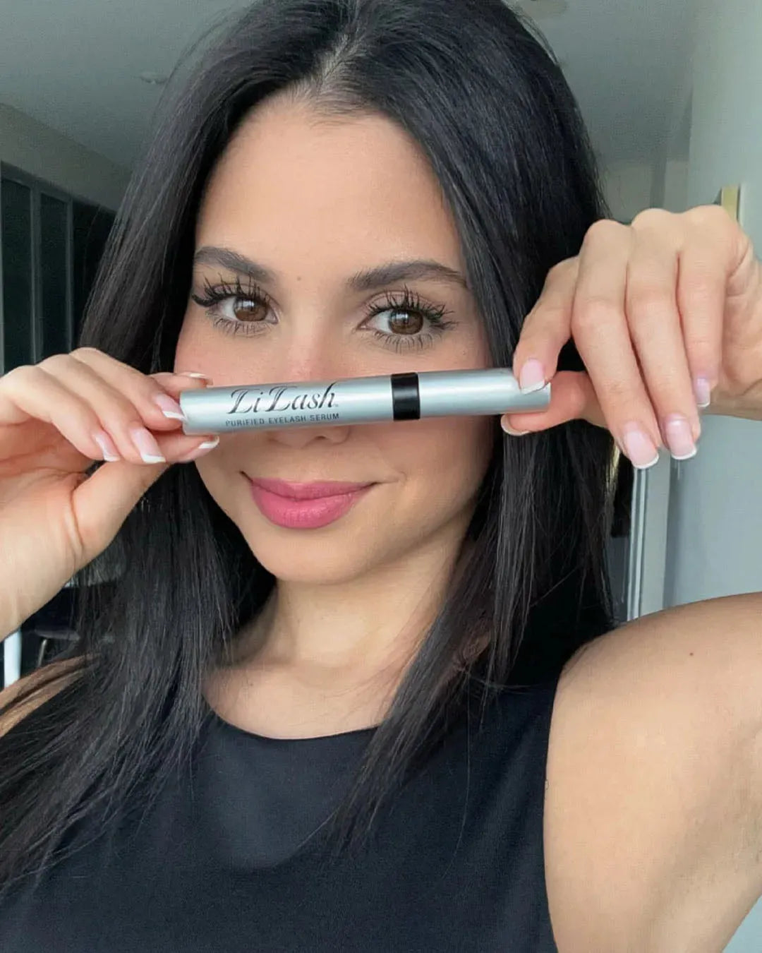 Woman holding LiLash purified eyelash serum, showcasing long, full lashes with natural makeup look
