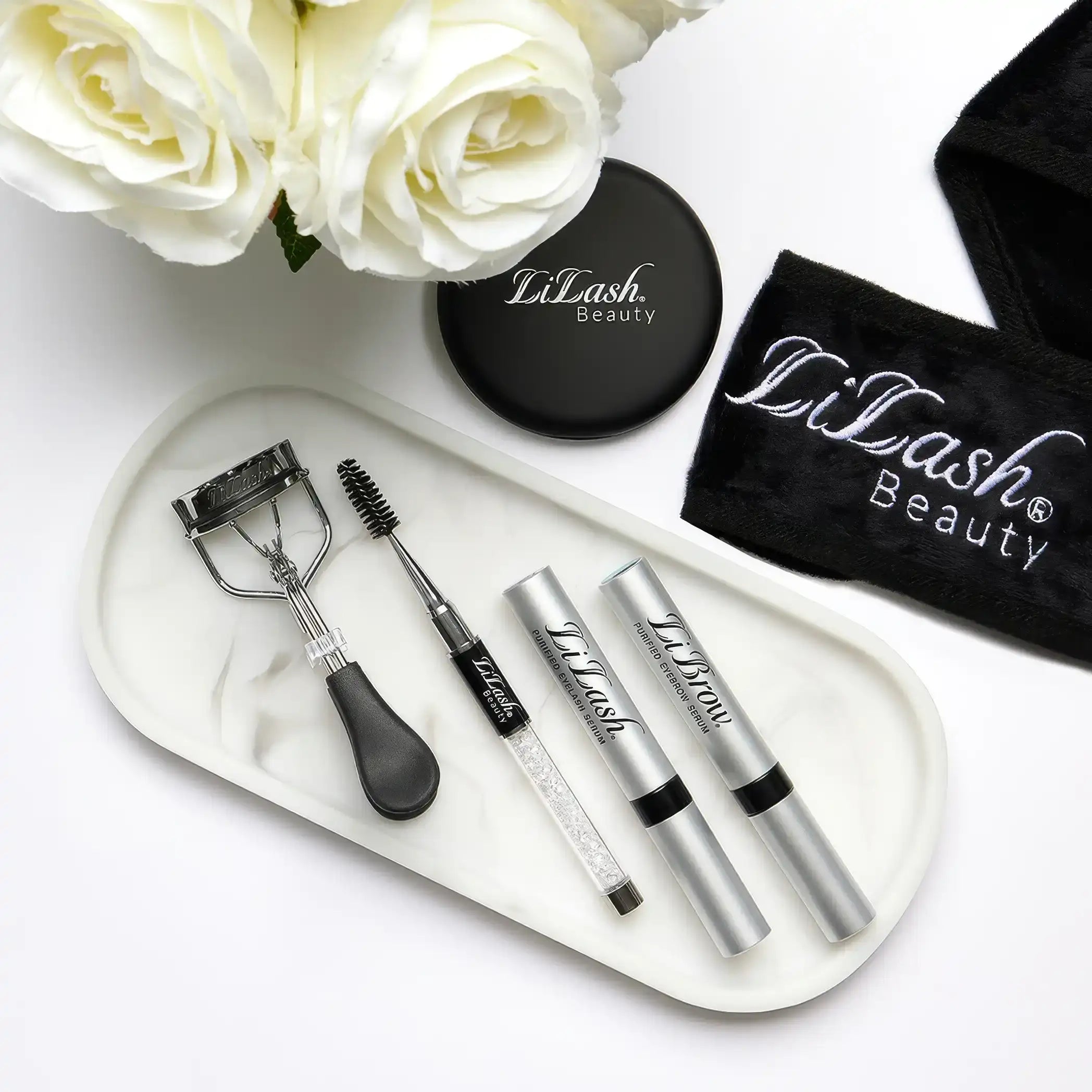 Eyelash and eyebrow serums with curler and spoolie on marble tray, white roses, black LiLash Beauty accessories