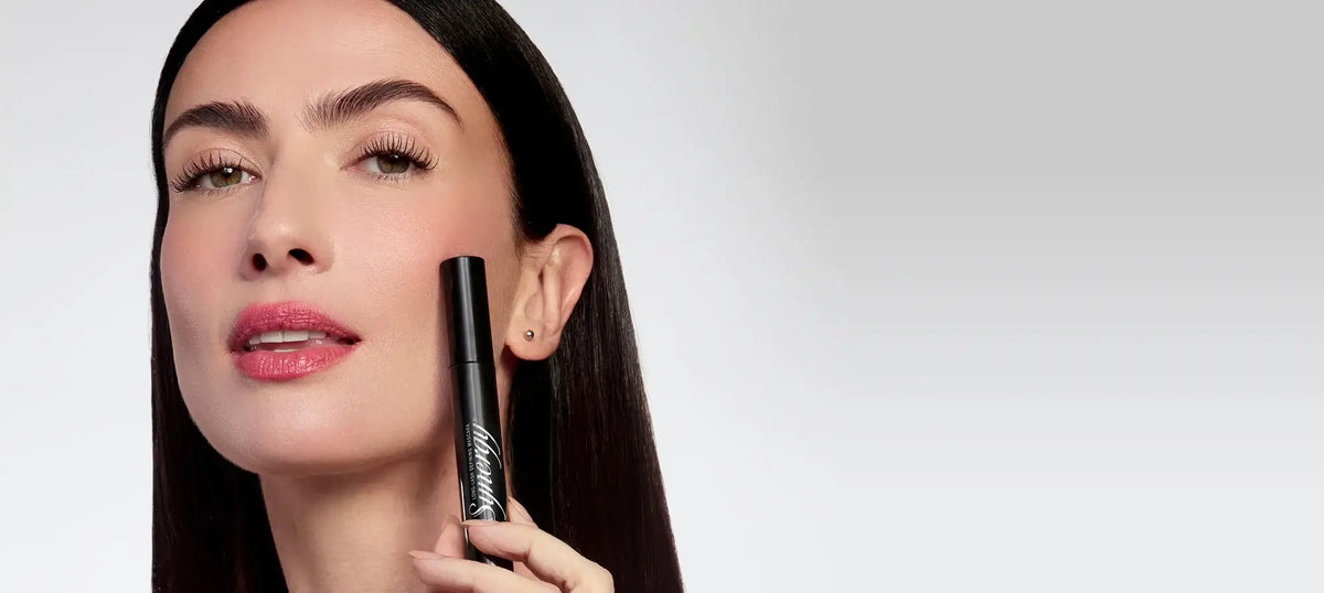 Woman holding a black LiLash eyelash serum tube next to her face.